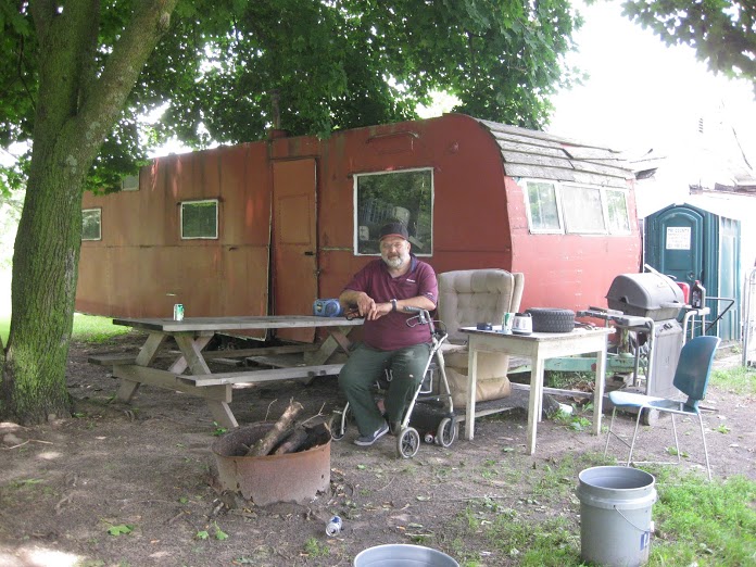 Robert Traviss sits on his walker in the yard of his Lake County home. The camper trailer behind him is where he keeps the tools he once used. He lives in another old trailer in the yard. (photo by Pat Shellenbarger)