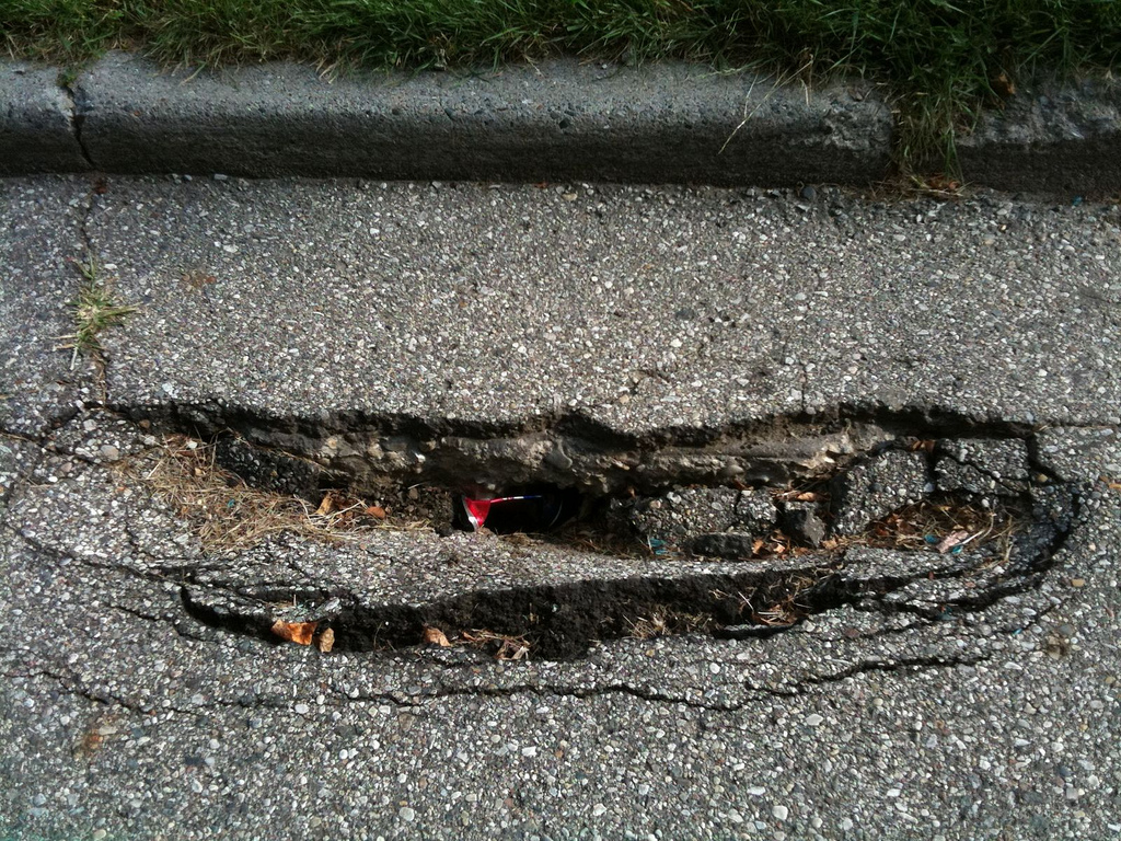 This pothole is deep enough to snag blown litter. Proposal 1, the May ballot question, seeks to raise money to fix it and thou thousands of others pocking Michigan roads. (Photo by the League of Michigan Bicyclists via Flickr; used under Creative Commons license)