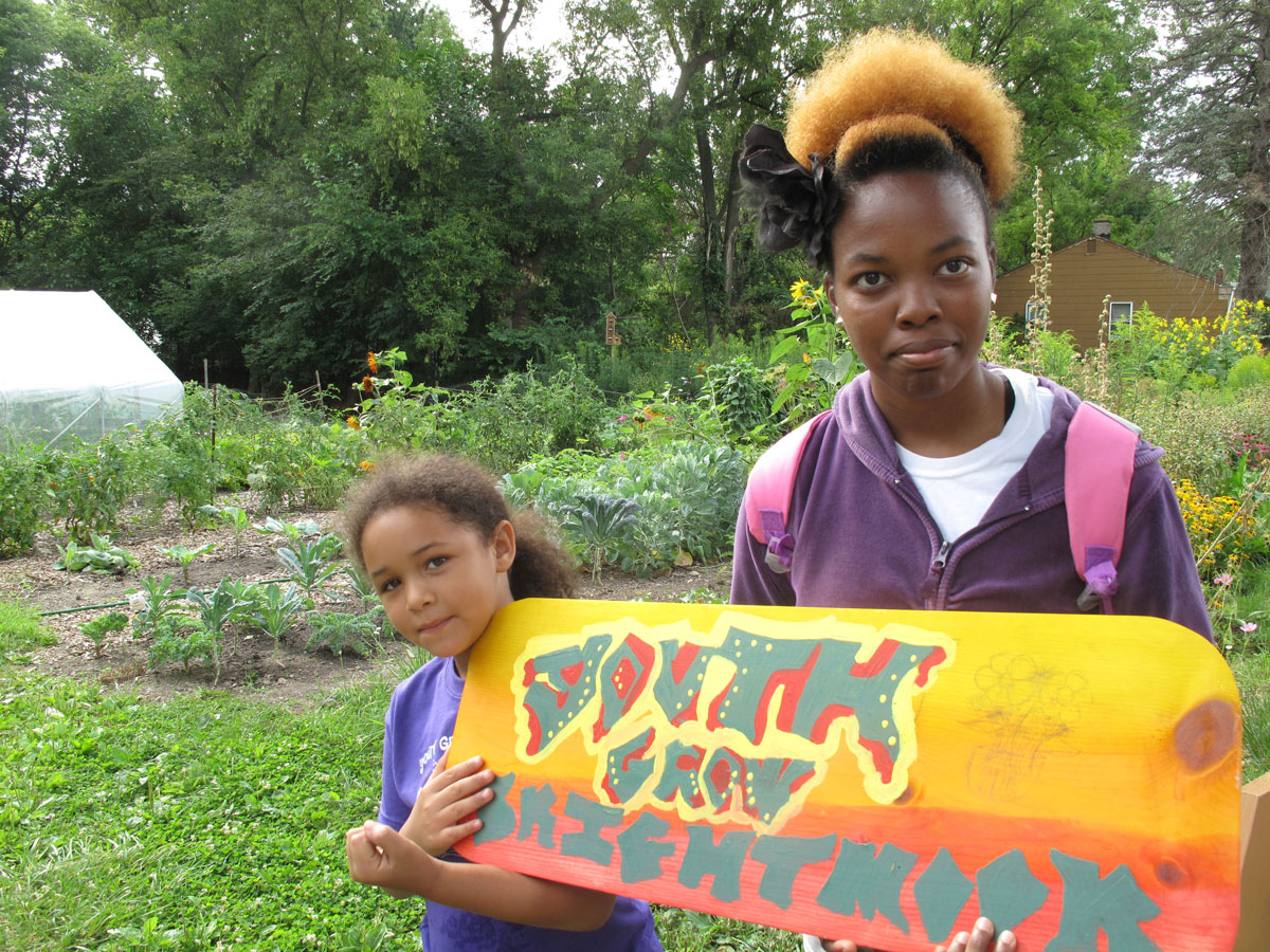 Nadia Dolphus, 7, and Shanna Bennett, 19, with A "Youth Grow Brightmoor" sign Shanna painted for one of numerous agricultural projects in the neighborhood. City officials see places like Brightmoor as pivotal to transforming Detroit's vacant land into productive assets. (Bridge photo by Bill McGraw)