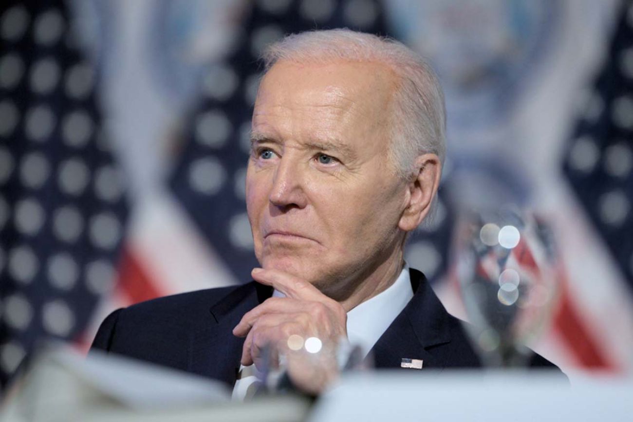 President Joe Biden with his hand on his chin. There are American flags in the background