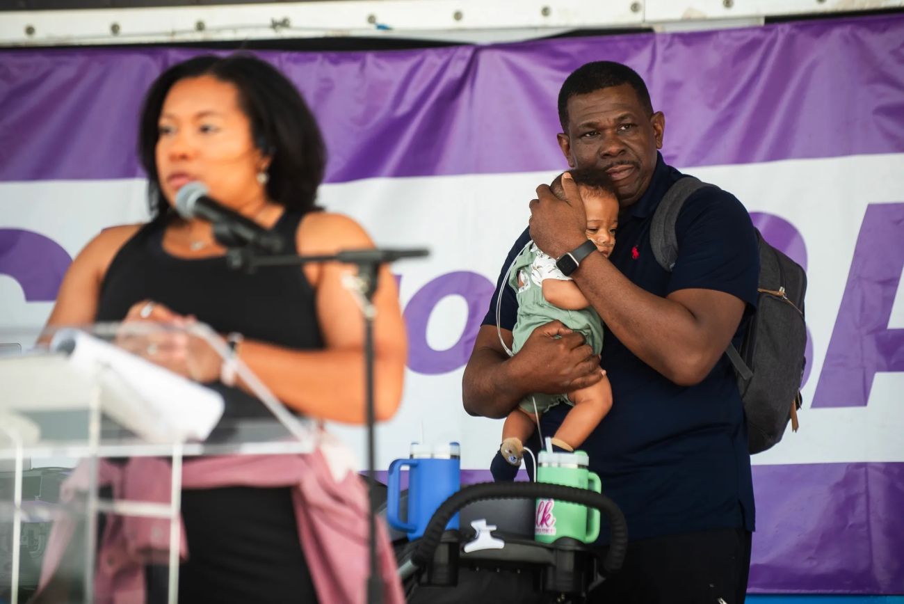 A woman speaks into a microphone on the foreground. A father holds a baby in the background