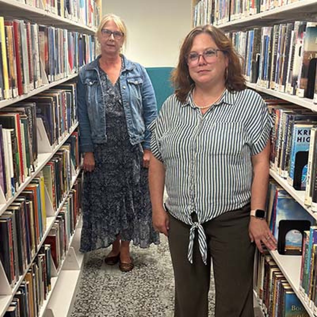 Alpena Library Assistant Director Jessica Luther and Director Debra Greenacre standing in between two bookshelves,