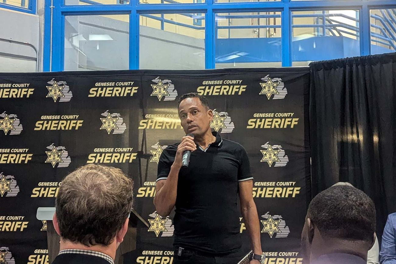 Hill Harper speaks into a microphone. Behind him is a sign for the Genesee County Sheriff's Office