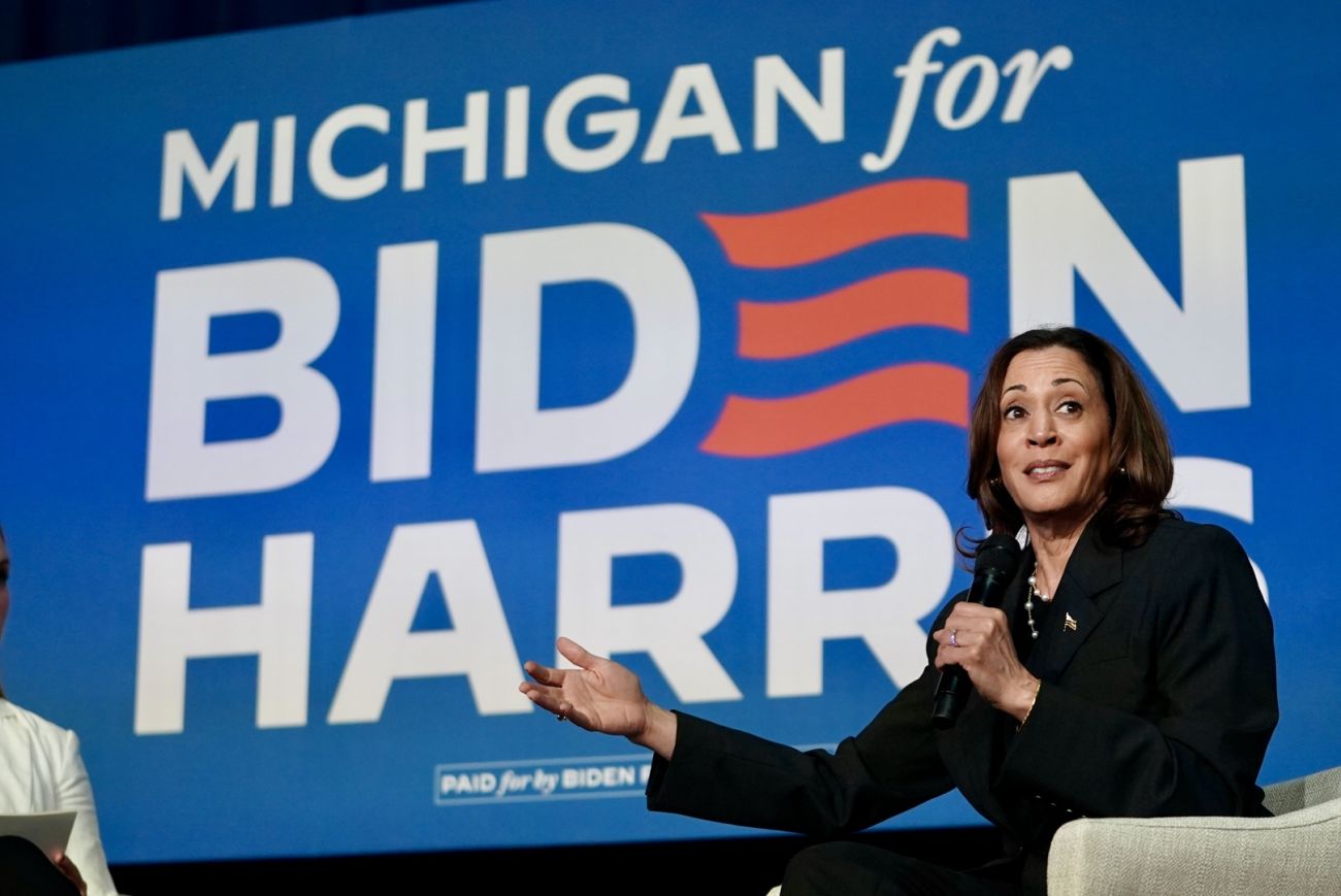 Vice President Kamala Harris sitting on a stage, wearing dark colored clothing. Behind her, a screen says "Michigan for Biden Harris"