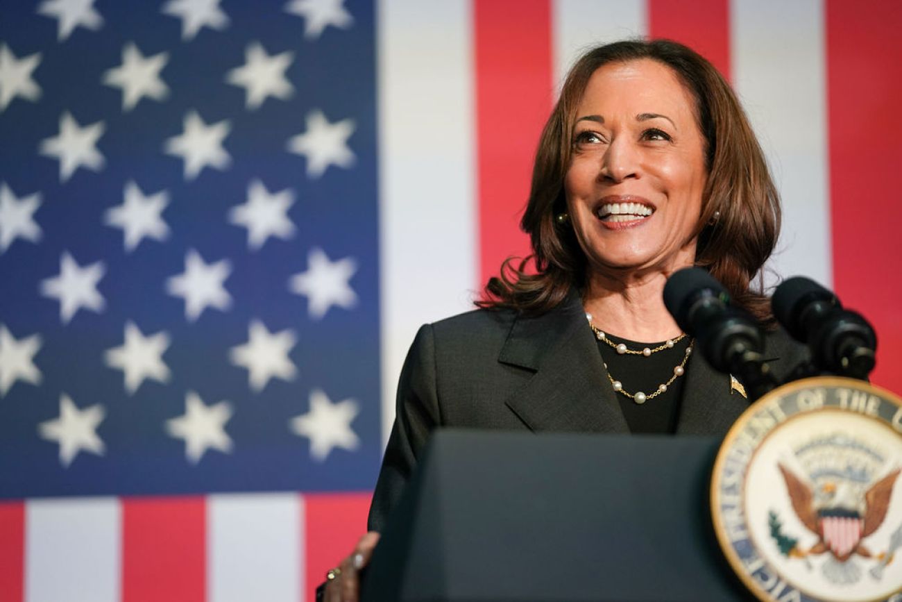 Vice President Kamala Harris, wearing dark-colored clothing, speaks at a podium. Behind her is the American flag