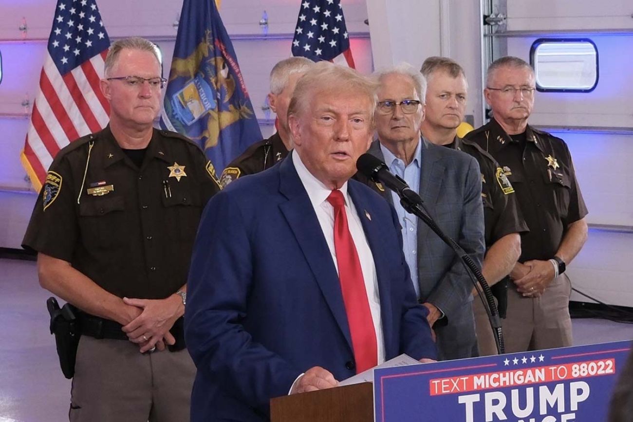 Donald Trump speaking into a microphone, law enforcement officers stand behind him