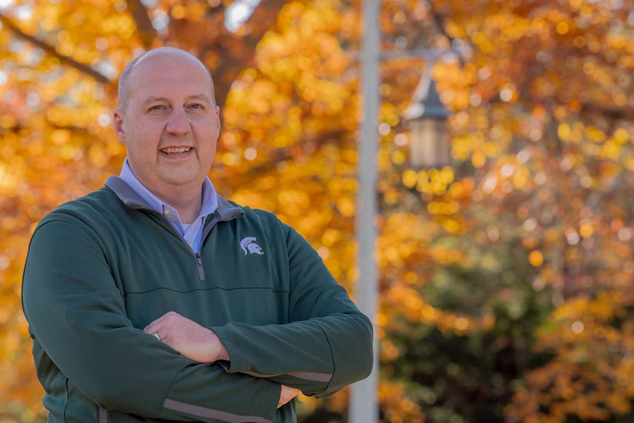 Democrat Curtis Hertel of East Lansing posing for a picture. He is wearing a green sweater and orange leaves behind him