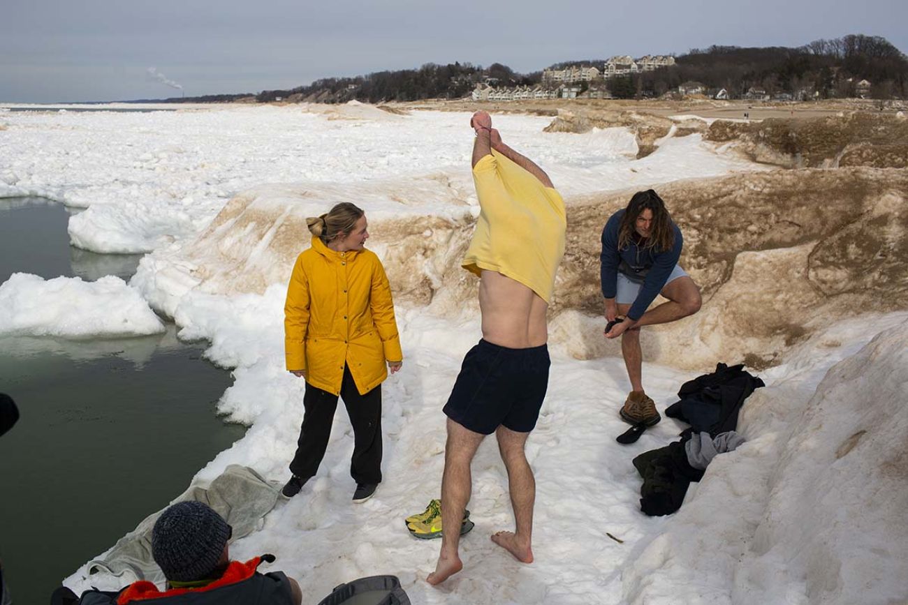 People in the snow, taking socks and shoes off. They are right by the edge of the water. 