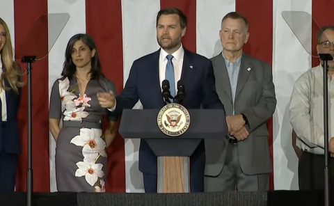 Vice President JD Vance standing in front of a podium. People standing next to him.