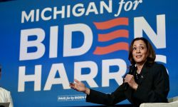 Vice President Kamala Harris sitting on a stage, wearing dark colored clothing. Behind her, a screen says "Michigan for Biden Harris"