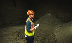 Hugo Staton, wearing an green vest and an orange hard hat, shines a flashlight on a pile of metal concentrate 