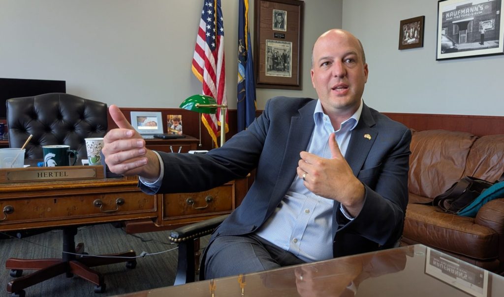 A man gestures while seated at a desk with an American flag in the background.