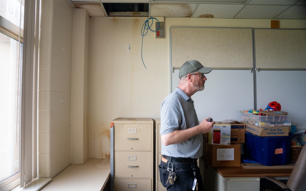 A man stands in a room at a school that's missing ceilling tiles and has water stains on ceiling.