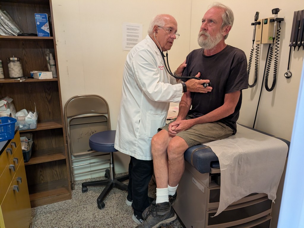 A male doctor uses a stethoscope on a male patient seated on n exam table