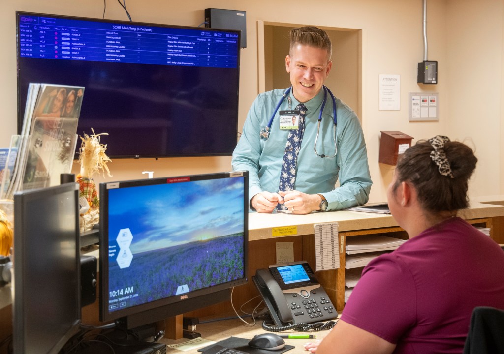 A smiling man leans on a counter in a hospital while chatting with a woman dressed in scrubs seated behind the desk.