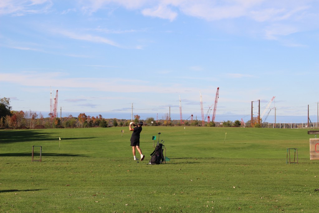 A person swings a golf club on a course with a construction site in the background.