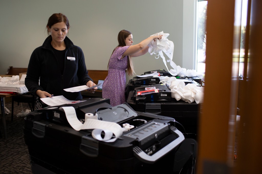 Two women work with voting machines during an election equipment test, examining paperwork and handling long strips of printed paper ballots.