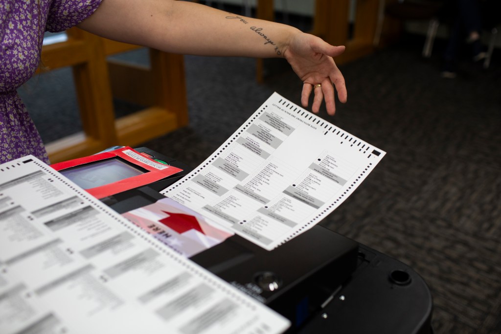 A person gestures toward a paper election ballot positioned above a voting machine during a demonstration of voting equipment.