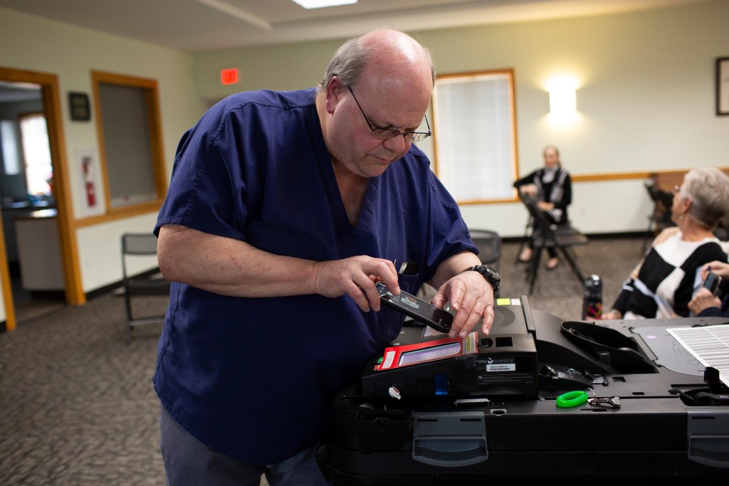 A man wearing navy blue scrubs and glasses examines voting equipment while holding a smartphone in a room with several seated people.