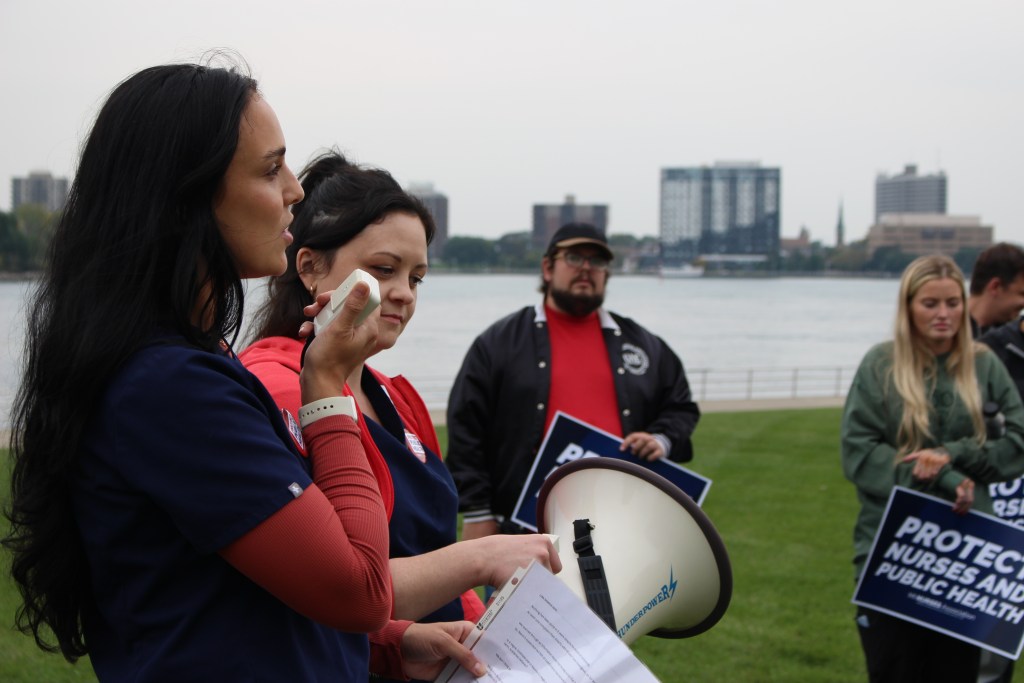 A nurse speaks into a megaphone at a waterfront rally, with attendees holding
