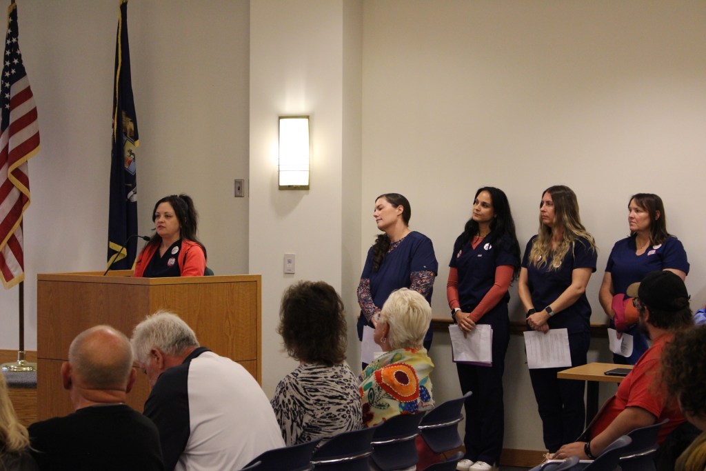 A woman speaks at a podium while four nurses in navy scrubs stand behind her holding papers, with audience members seated in the foreground.